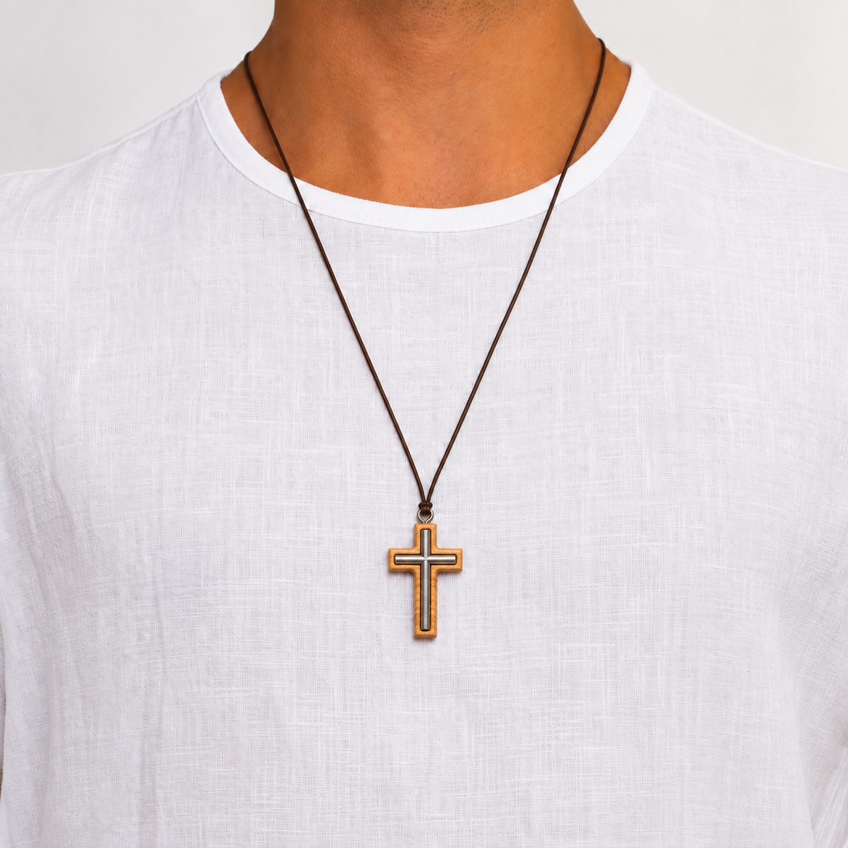 A close-up of a person wearing a brown cord necklace with a gold cross pendant, styled with a white linen t-shirt against a light background.