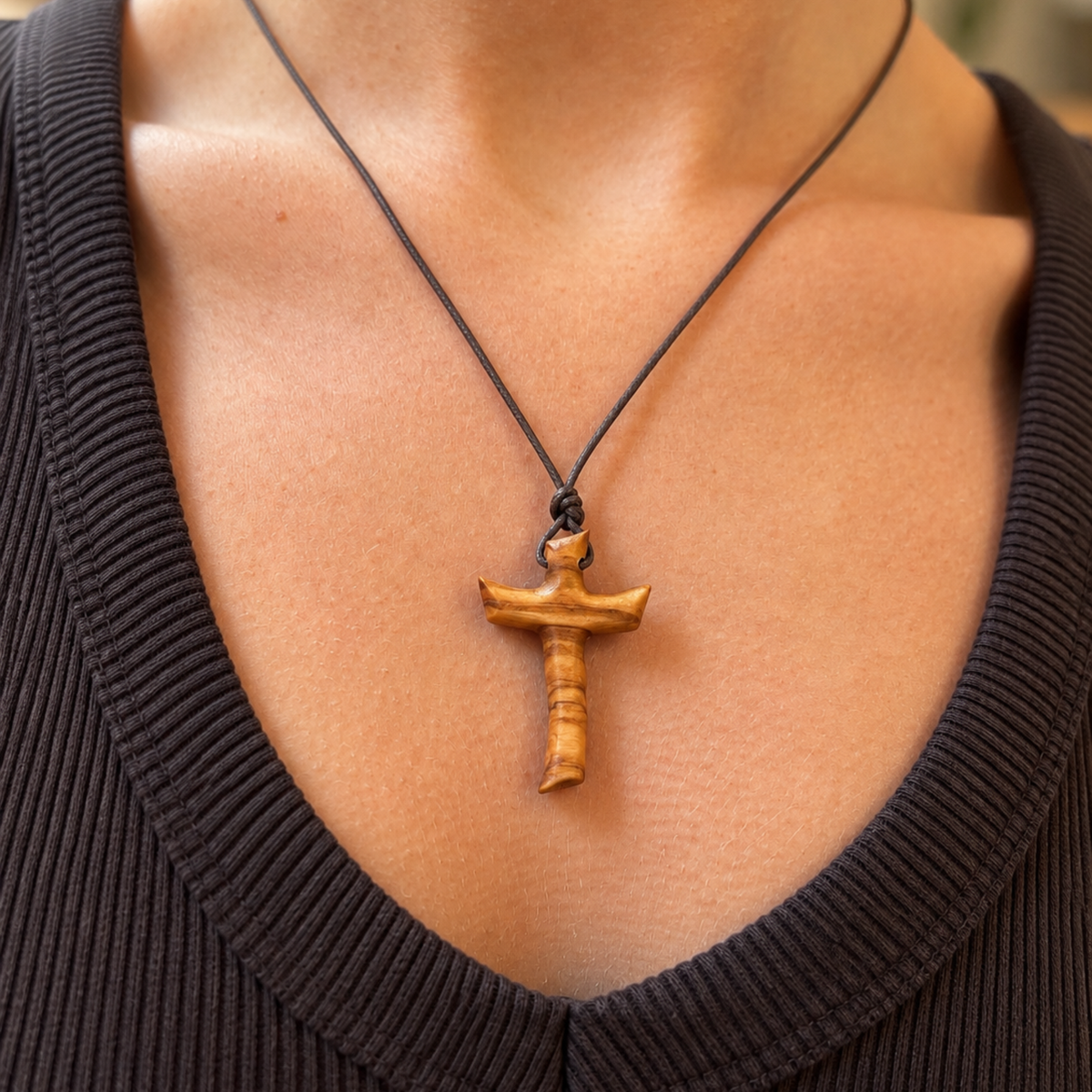 A close-up of a person's chest wearing a black cord necklace with a carved wooden cross pendant, paired with a dark ribbed V-neck top.